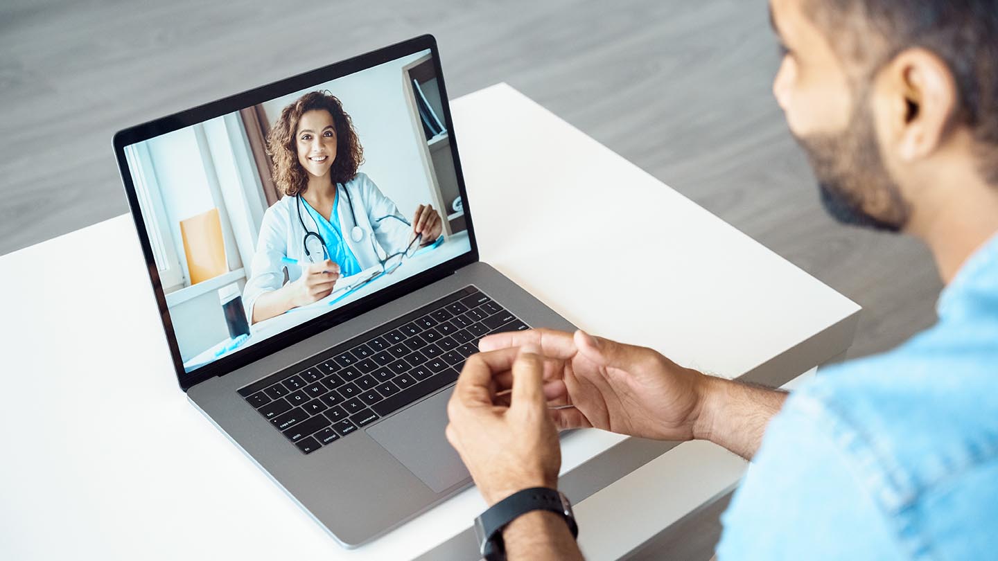 View over male client shoulder sit at desk receive medical consultation online from female doctor. Indian man listening practitioner. Distant communication, health protection and telemedicine concept