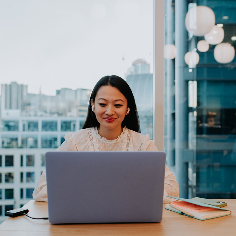 A young woman in a modern office space uses a laptop at a desk. Space for copy.