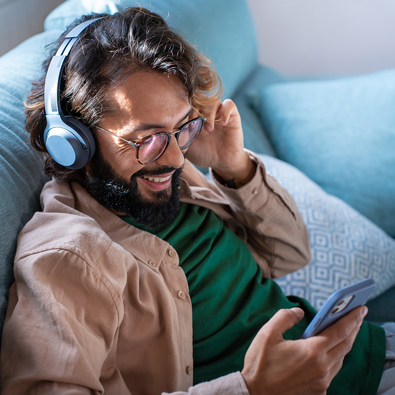 Smiling young man wearing headphones listen music playing in smartphone sitting on sofa at home