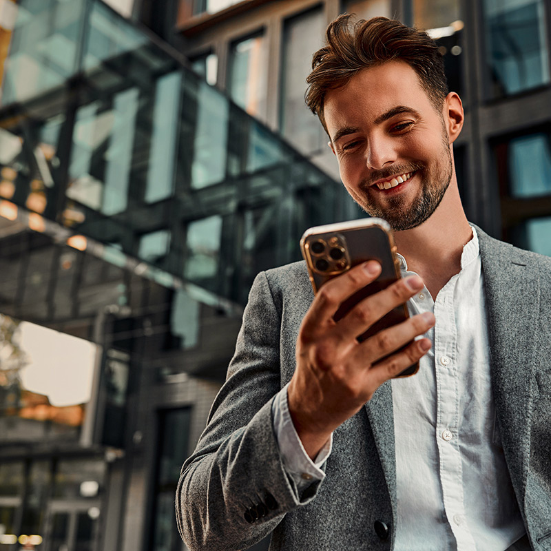 Portrait of attractive confident modern smiling happy adult dressed gray suit man holding a phone and standing outside near office building. Online work, communication. Copy space.