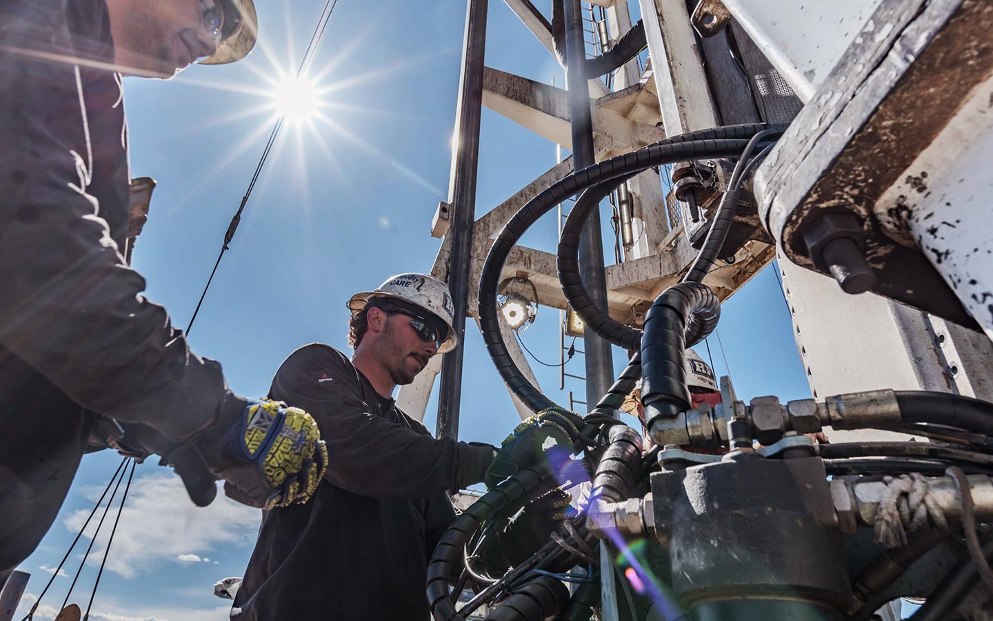 Two workers in hard hats and safety glasses adjust hoses and fittings on drilling equipment in bright sunlight, with the sun flaring in a clear blue sky.