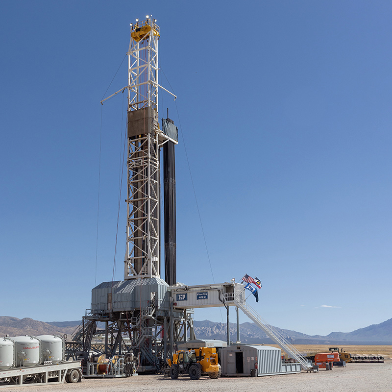Tall drilling rig on a flat, open site with equipment and vehicles at the base, set against distant mountains and a clear blue sky.