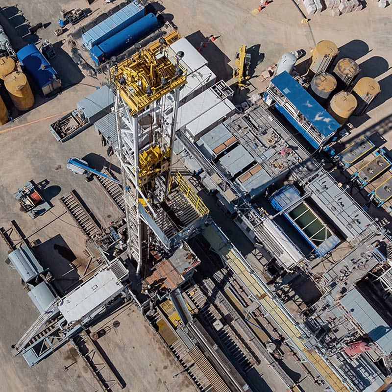 Top-down aerial view of a drilling site with a tall rig at center, surrounded by equipment, tanks, and service vehicles arranged on a dusty work pad.