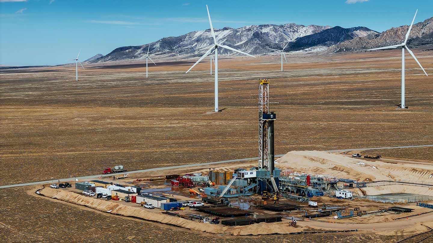 Drilling site in a wide, open plain with wind turbines nearby and snow-dusted mountains in the background under a clear blue sky.