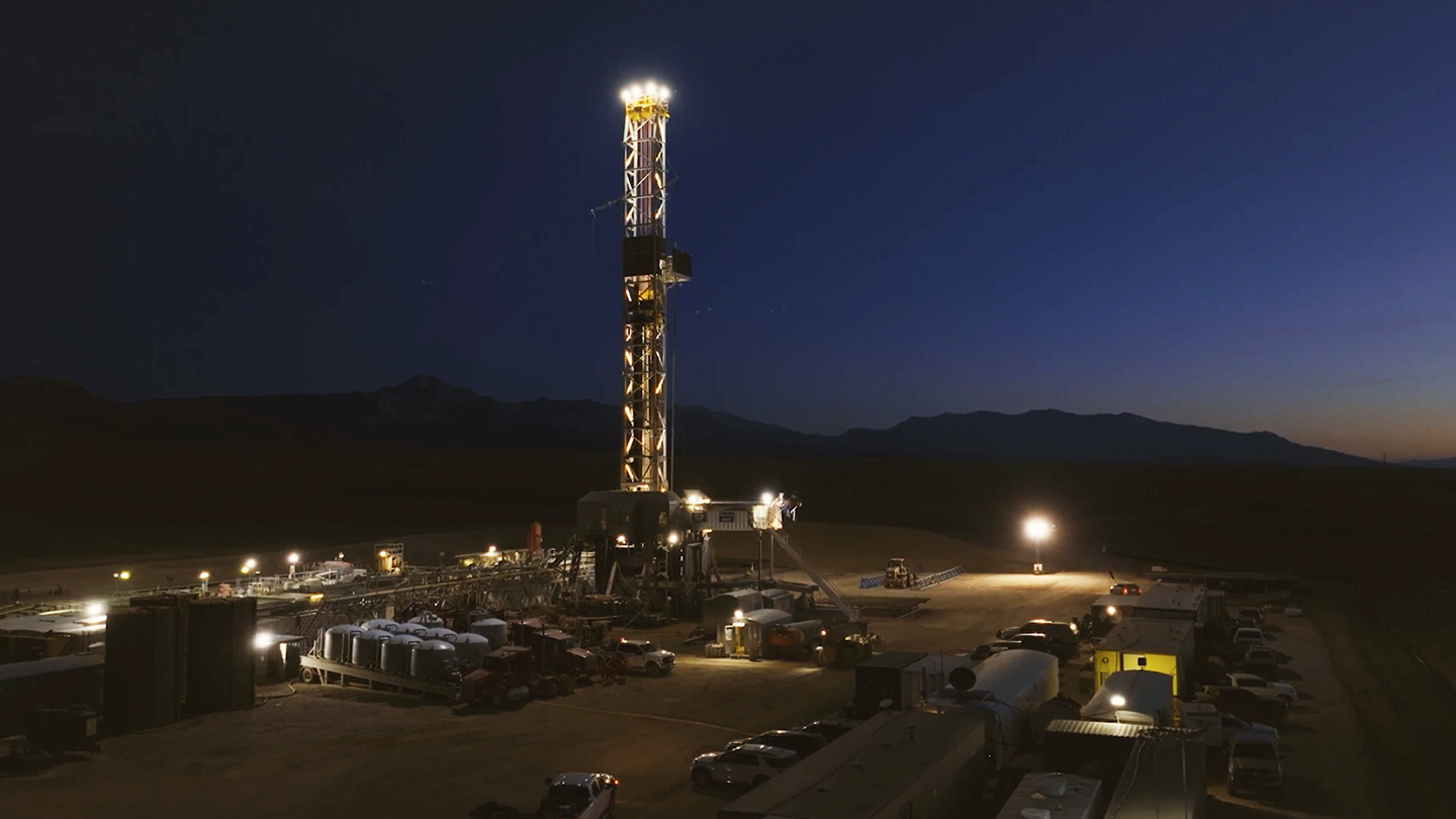 Floodlit drilling rig and worksite at night, with parked vehicles and equipment in the foreground and dark mountain silhouettes under a deep blue sky.