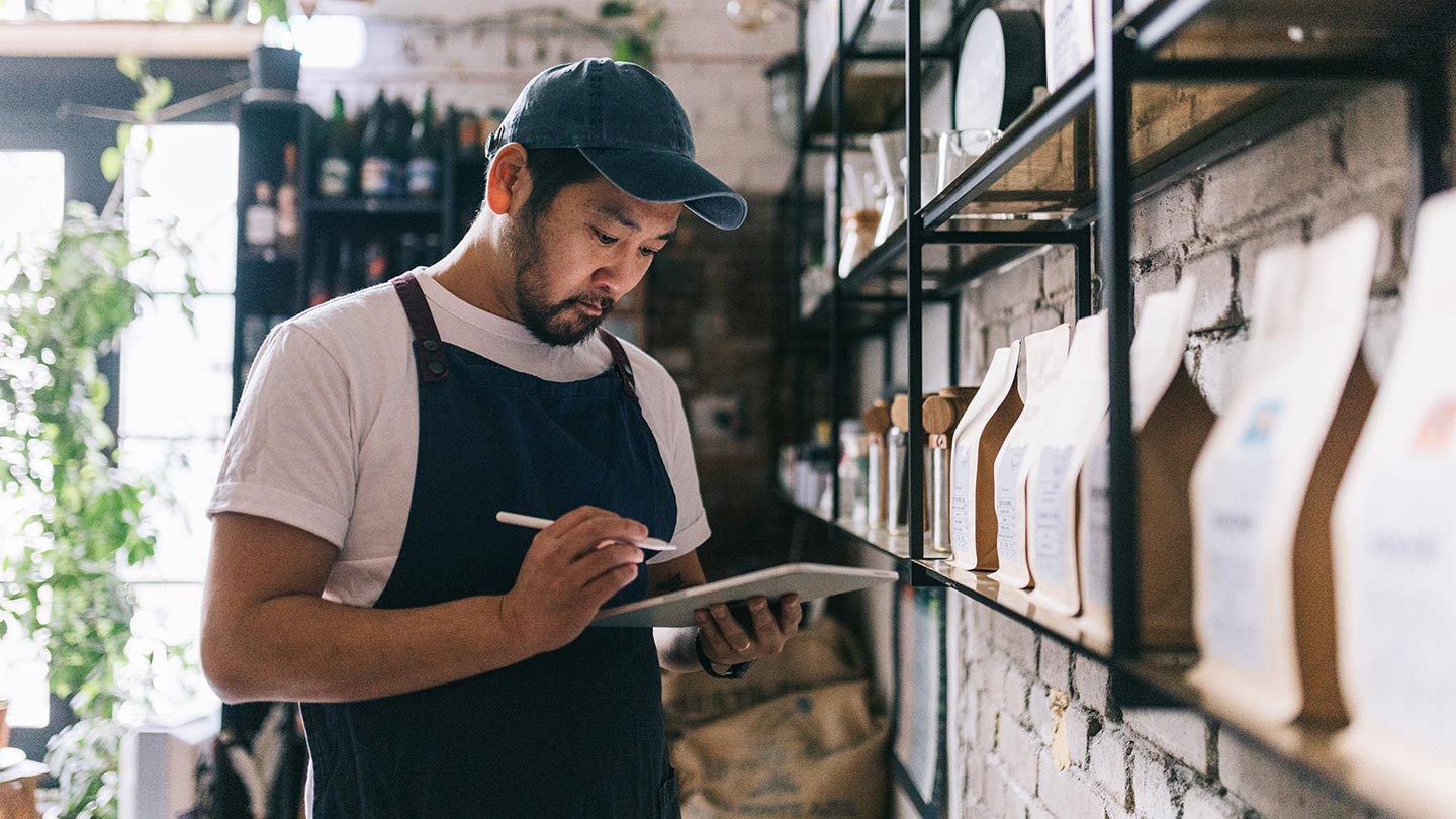 Person wearing an apron and cap taking inventory with a tablet in a small shop, standing beside shelves of packaged products.