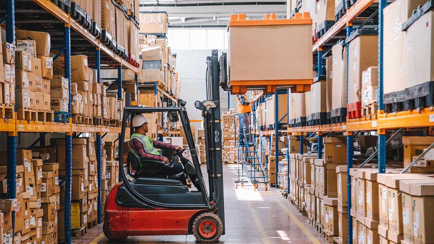 A warehouse aisle with tall shelves of boxed inventory, where a forklift operator is lifting a large pallet/bin while another worker stands on a ladder in the background.