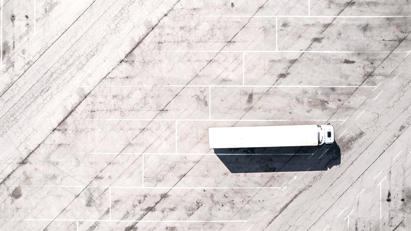 Overhead view of a lone white semi‑truck parked on a large, empty, cracked concrete lot with long shadows.