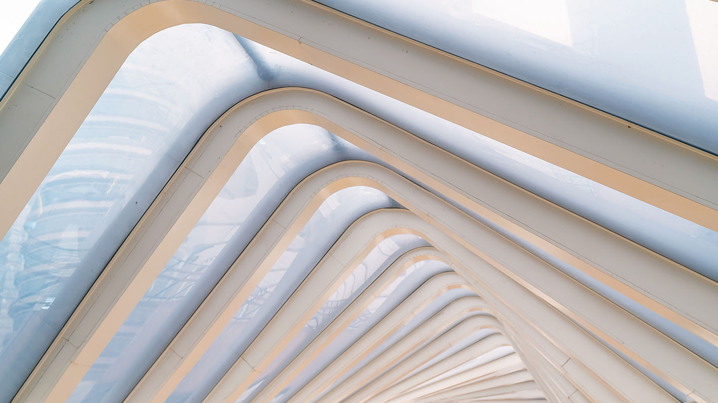 Angled view of repeating white steel arches under a translucent skylight, creating a geometric, tunnel‑like pattern.