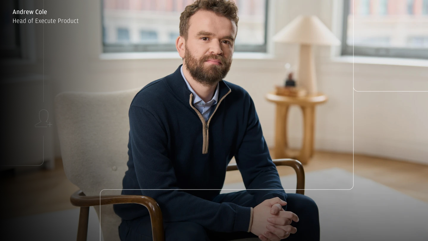 Portrait of a bearded man seated in an armchair with hands clasped, in a bright room with large windows and a side table lamp; faint UI-style overlay lines and a name/title caption appear in the upper-left corner.