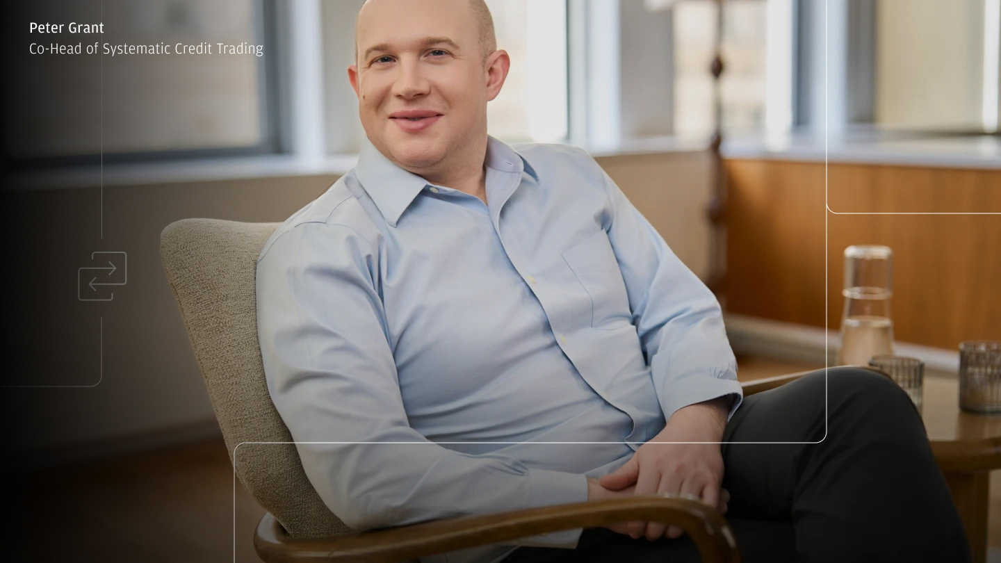 Portrait of a smiling man in a light blue dress shirt seated in an armchair in a bright office lounge, with a small table holding a water carafe and glasses in the background; subtle interface-style overlay lines/icons and on-screen text reading “Peter Grant — Co-Head of Systematic Credit Trading.”