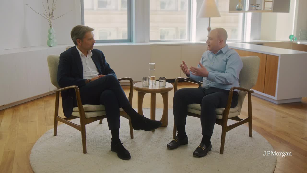 Two people in business attire sit in armchairs facing each other in a bright office lounge, talking beside a small round table with a water carafe and two glasses; “J.P. Morgan” watermark in the lower-right.