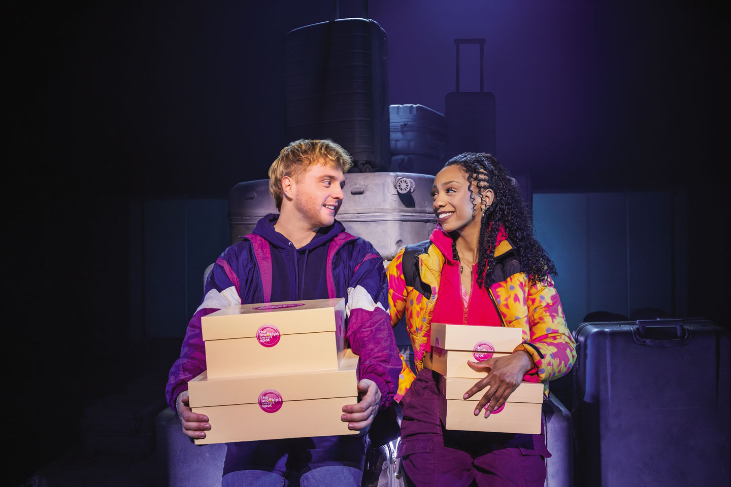 Two people in colorful jackets sit on stage holding cake boxes, with suitcases behind them.