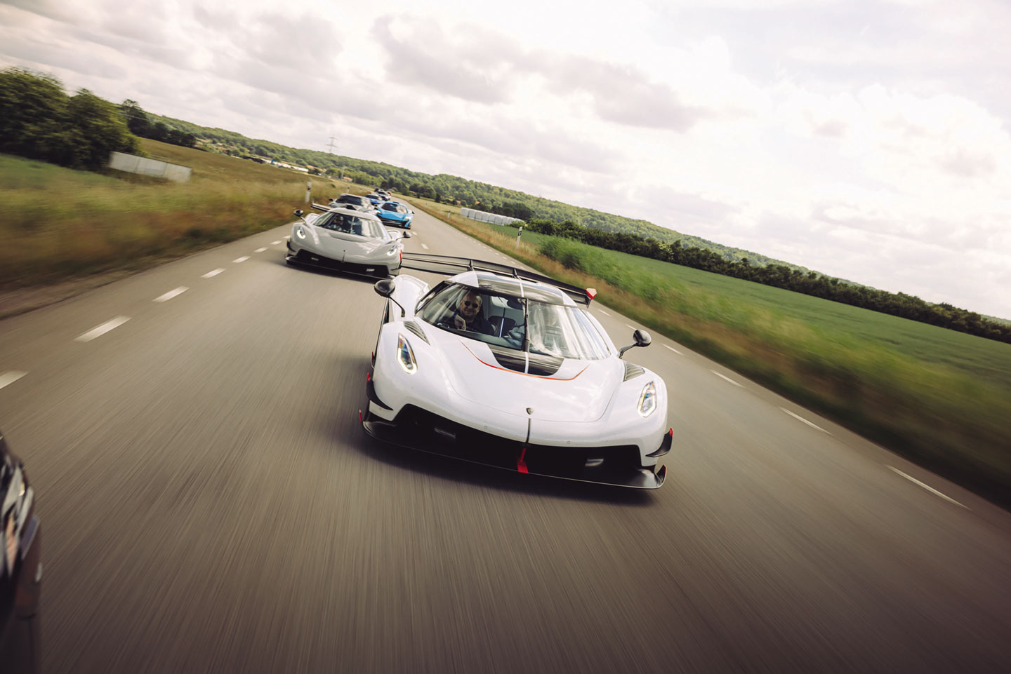 A group of high-performance sports cars drives down a scenic road.